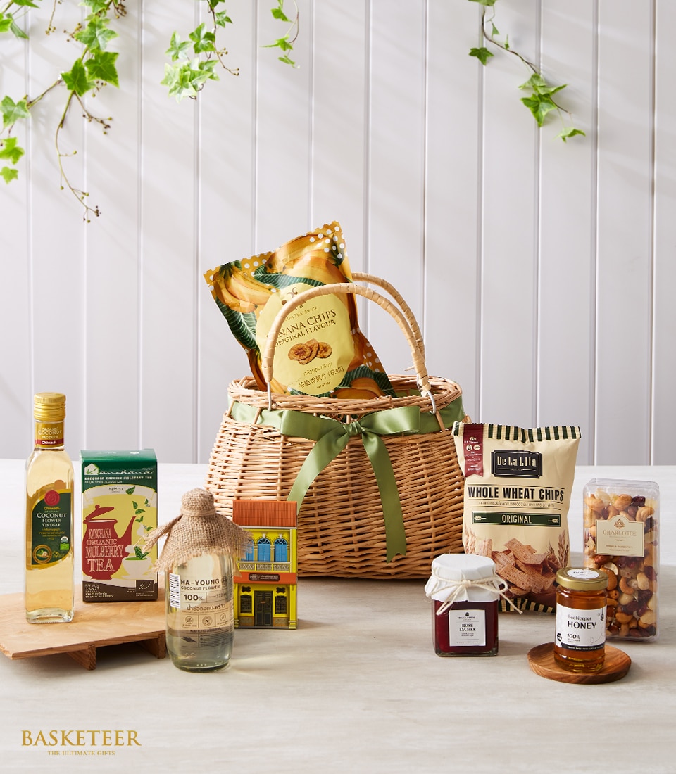 A luxurious assortment of gourmet delights tastefully arranged beside an elegant wicker basket adorned with a sophisticated green ribbon, proudly showcasing the Basketeer logo. This indulgent collection features artisanal chips, premium olive oil, exquisite tea, rich honey, gourmet crackers, and a charming house-shaped gift box accompanied by delectable jam. The scene is set against a light vertical striped background with lush green leaves gracefully hanging above, creating an inviting ambiance.
