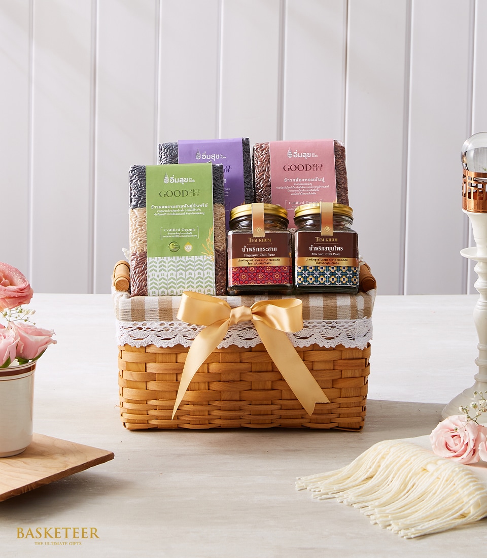 A luxurious wicker gift basket adorned with an elegant beige bow sits enticingly on a chic beige table, accentuated by a serene light-colored, vertical paneled wall. The exquisite basket, boasting the distinguished Basketeer logo on a refined ribbon, cradles an array of vibrant fabric goods and artisanal glass jars. Nearby, a vase of delicate pink flowers exudes freshness and romance alongside a plush cream-colored fringed cloth perfectly folded to complete this sophisticated and inviting scene.