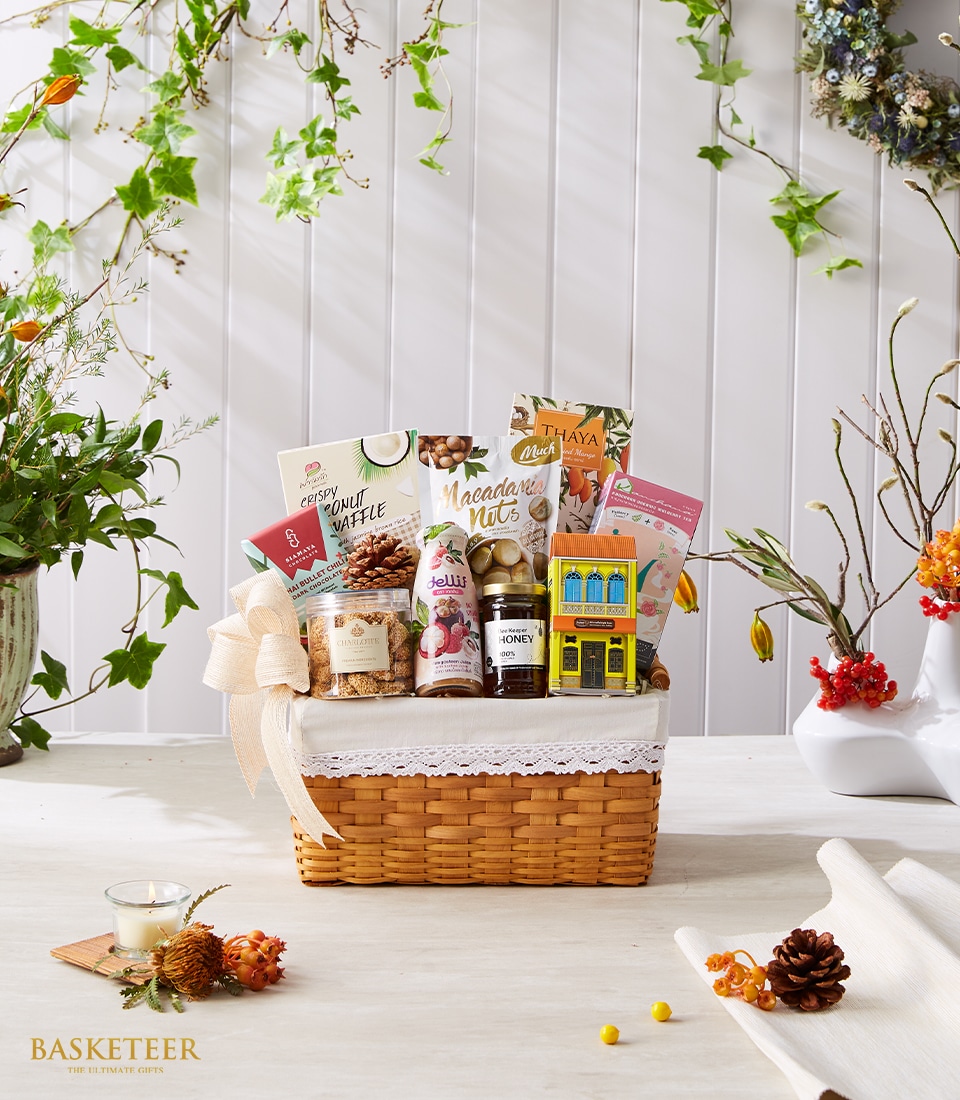 An elegant wicker basket, curated by Basketeer, rests gracefully on a wooden table. Overflowing with an exquisite selection of premium snacks, artisan honey, freshly pressed juice, and indulgent bath salts, this basket exudes luxury. The white lace ribbon that embellishes the basket adds a touch of sophistication. Encircled by charming autumn-themed decorations—pine cones and delicate blossoms—the scene is both inviting and opulent. A distinguished 