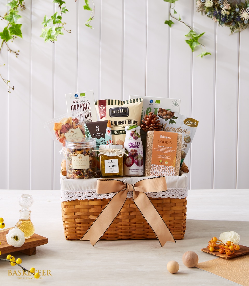 A gorgeously curated Basketeer gift basket brimming with an exquisite selection of organic snacks and gourmet delights, including artisan popcorn, luscious dried fruits, premium nuts, and savory crackers. Adorned with an elegant tan bow featuring the Basketeer logo, this luxurious offering is set atop a light wooden table graced with tasteful decorative accents.