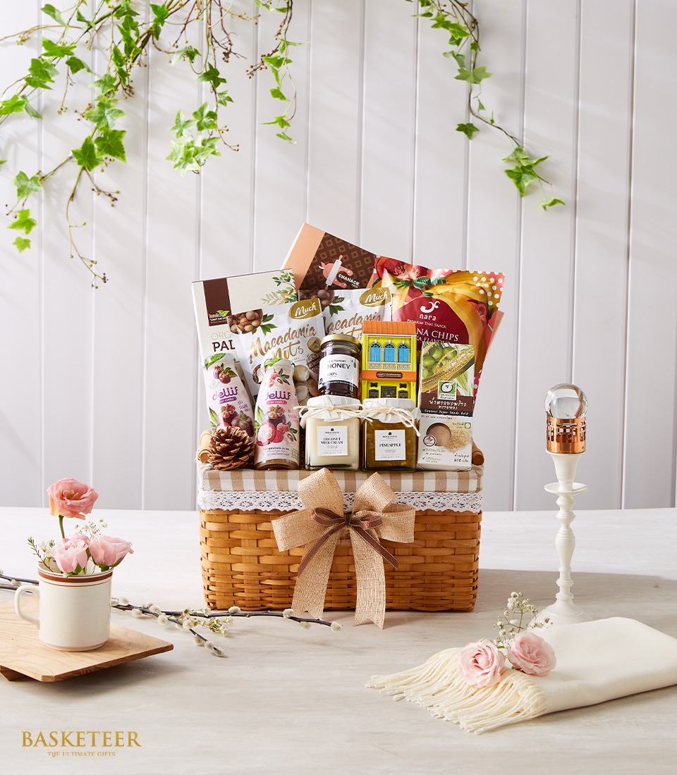 An exquisite woven gift basket brimming with an assortment of luxurious treats, including decadent chocolates, artisanal nuts, and gourmet jars, all elegantly wrapped with a beige ribbon showcasing the Basketeer logo. This sophisticated arrangement rests on a beautifully set table adorned with delicate flowers and an ornate candlestick atop a vintage lace cloth. The scene is framed against a pristine white wooden backdrop intertwined with lush green vines, creating an inviting atmosphere of opulence and refinement.