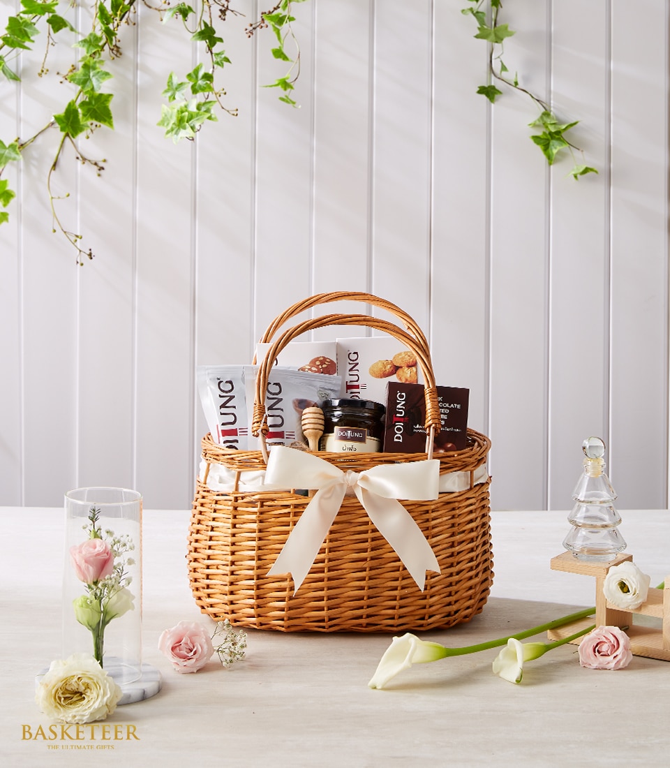 In an opulent wicker basket accented with an elegant white ribbon displaying the Basketeer logo, a curated selection of gourmet snacks and a charming jar beckons indulgence. The scene is adorned with a glass vase filled with exquisite blooms, a delicate glass dome showcasing a single blush pink rose, and an artisanal clear Christmas tree figurine for a touch of festive sophistication. Verdant leaves drape gracefully in the background, completing this luxurious and inviting tableau by Basketeer.