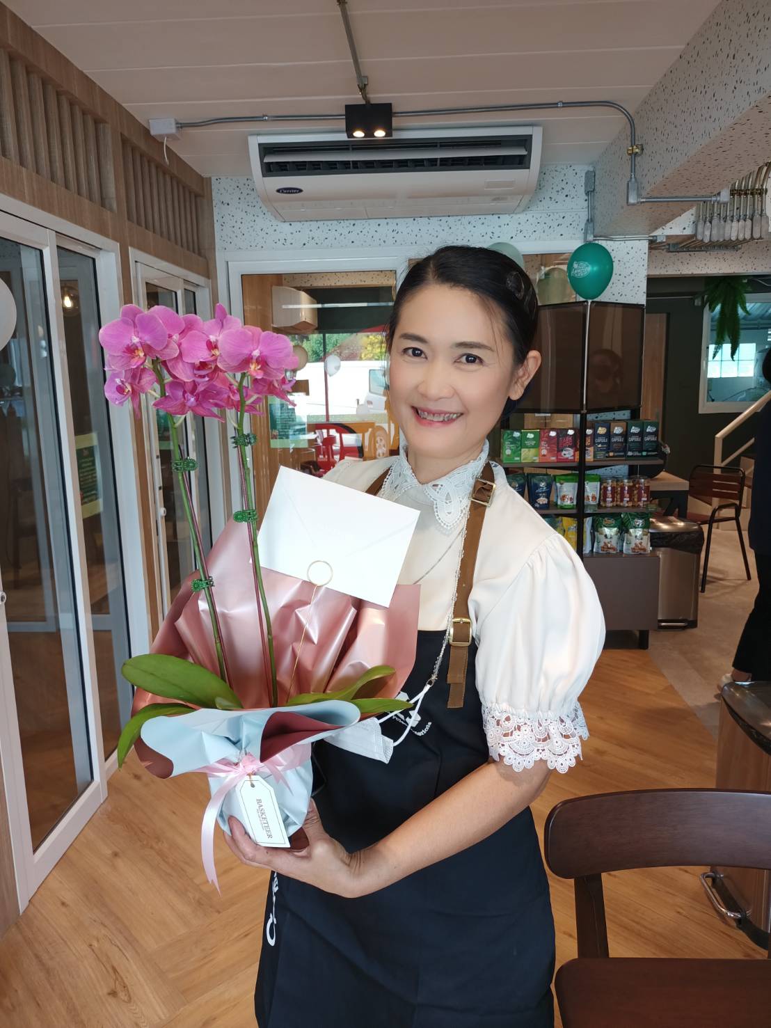 A smiling woman, dressed in a black apron over a white blouse with lace sleeves, holds a bouquet of pink orchids and a small white envelope. She stands in what appears to be a cafe with wooden floors and glass doors in the background.