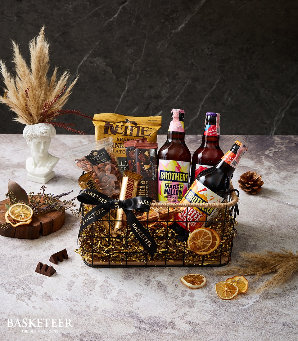 A lavish wire basket brimming with an exquisite selection of gourmet snacks and beverages. Among the treasures inside, you'll find elegant bottles of premium drinks, luxurious chocolate bars, and delectable snack packs. Accentuating this sumptuous assortment are dried orange slices and sophisticated rustic decorations. The scene is graced by delicate pampas grass set against a rich, dark textured backdrop—an opulent display crowned with the signature Basketeer ribbon and logo-adorned box, embodying pure indulgence.