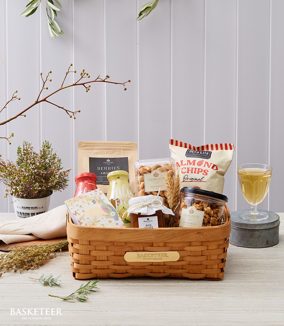 A luxurious wicker basket, elegantly curated by Basketeer, graces the polished wooden table. It overflows with gourmet delights: a bag of artisan almond chips, a jar brimming with premium mixed nuts, and an exquisitely packaged box labeled 