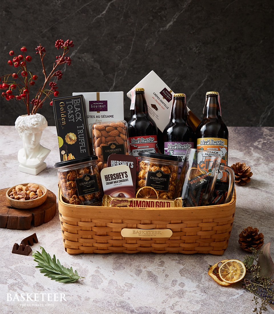 A luxurious wicker gift basket from Basketeer boasts an exquisite selection of gourmet delights and artisanal beverages. The opulent assortment includes rich chocolates, premium almonds, savory sesame croquets, and elegantly bottled root beer, complemented by a captivating variety of indulgent snacks. Nestled amidst elegant props like a refined small bust and a delicately dried orange slice on a polished stone surface, this scene exudes sophistication and invites you to savor the lavish offerings within. The basket is adorned with a tasteful ribbon featuring the prestigious Basketeer logo, enhancing its sumptuous allure.