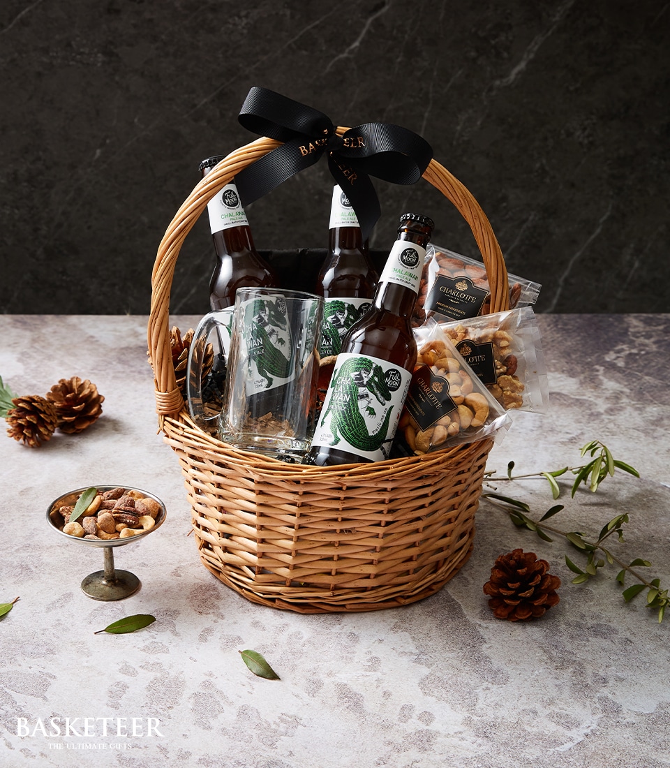 A luxurious wicker gift basket overflowing with premium bottles of craft beer, a finely crafted glass, gourmet packets of nuts, and artisanal snacks. The basket is elegantly adorned with a black ribbon featuring the distinguished Basketeer logo around the handle. Nestled within a serene setting of textured surfaces, pine cones, and lush greenery—this scene encapsulates the essence of indulgence and sophistication by Basketeer.