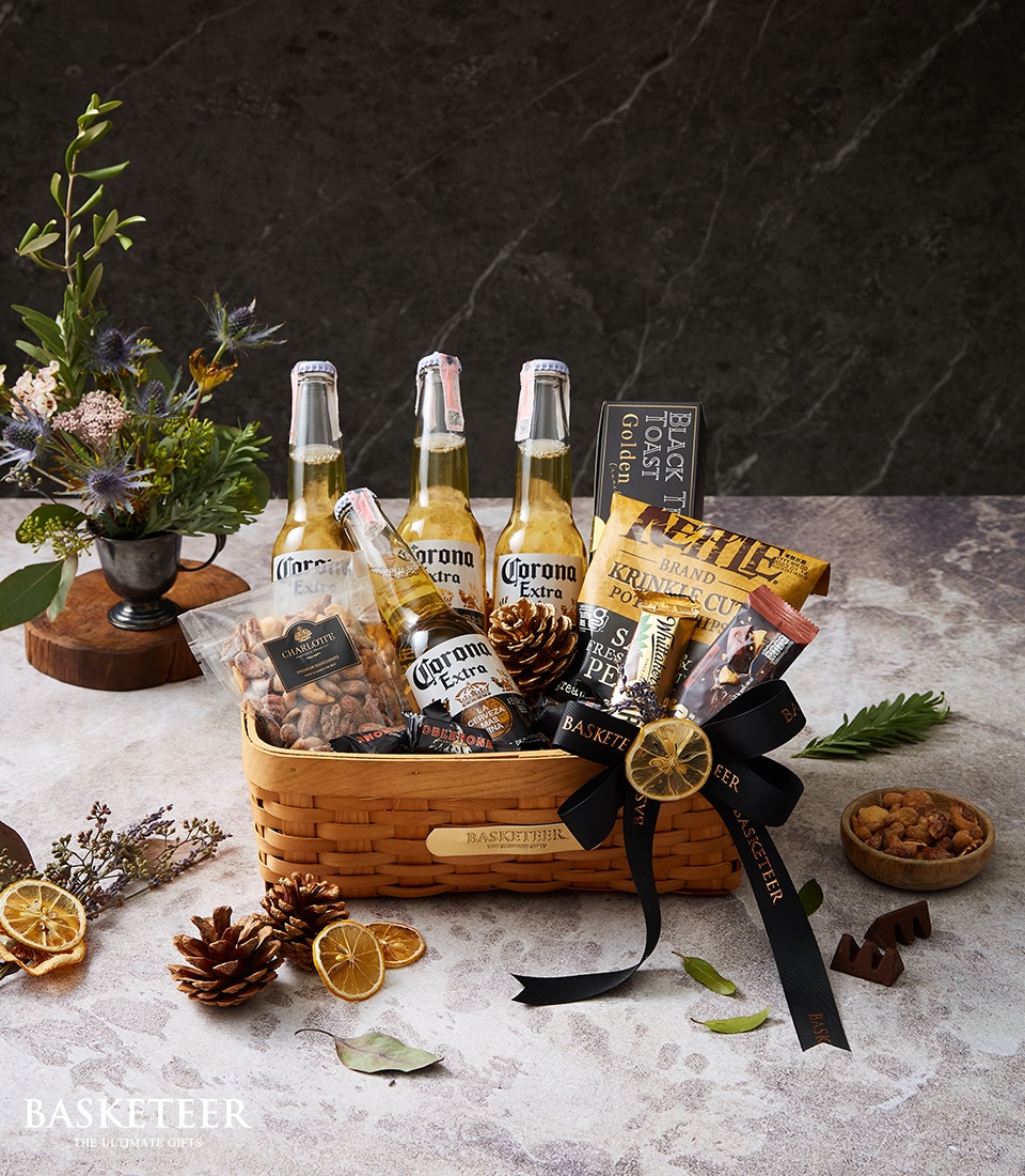 A luxurious gift basket featuring Corona beers and an array of gourmet snacks, adorned with a tasteful decorative black ribbon bearing the Basketeer logo. Inside, an elegant small box labeled 