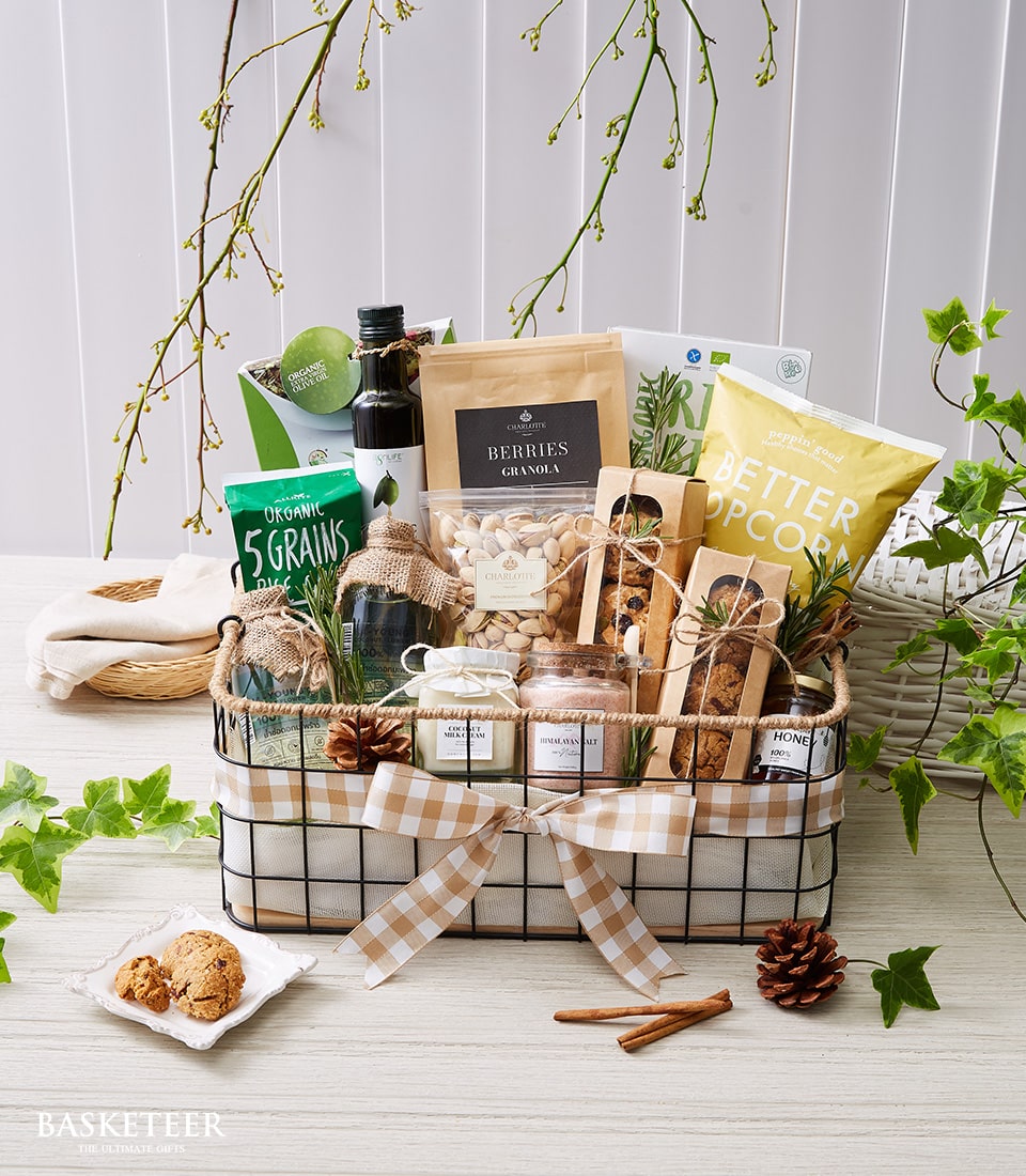 กระเช้าของขวัญสุดหรูจาก Basketeer ประดับด้วยริบบิ้นสีเบจหรูหราที่มีโลโก้ Basketeer แสดงถึงเมนูรสเลิศที่ได้รับการคัดสรร รายการต่างๆ ที่คัดสรรมาอย่างดีบรรจุอยู่ในภาชนะลายตารางหมากรุกที่ประดิษฐ์อย่างสวยงาม ได้แก่ น้ำมันมะกอกระดับพรีเมียม กราโนล่าสูตรพิเศษ ป๊อปคอร์นรสเลิศ ถั่วผสมรสเผ็ด มะเดื่อเนื้อฉ่ำ แครกเกอร์ชั้นดี และมะม่วงทอดกรอบที่แปลกใหม่ ตะกร้าเปล่งประกายความซับซ้อนและล้อมรอบด้วยความเขียวขจีและโคนต้นสนแบบชนบท ดื่มด่ำไปกับความหรูหราของเครื่องบูชาของ Basketeer ขณะที่เปลี่ยนฉากต่างๆ ให้กลายเป็นฉากแห่งความสง่างามที่ประณีต