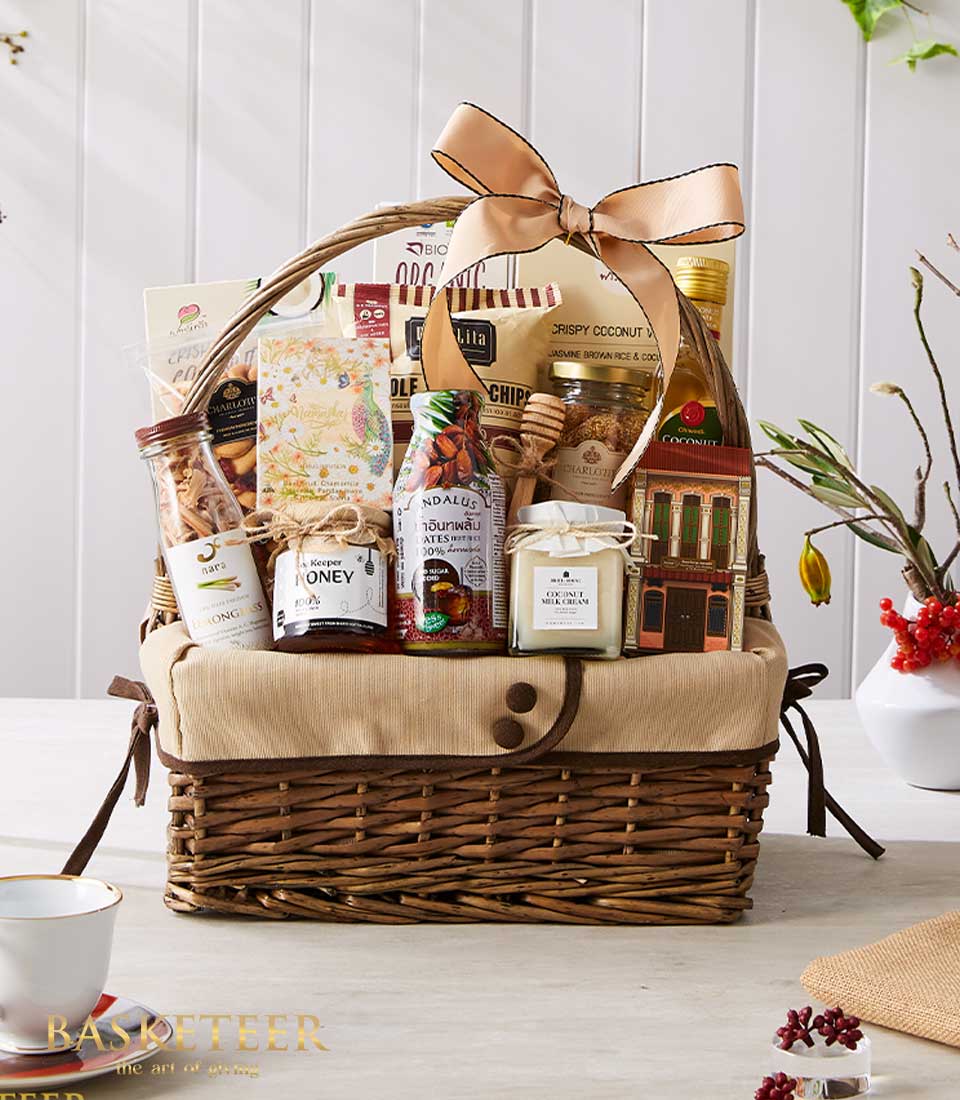A sumptuous wicker basket, delicately wrapped with an elegant beige cloth and adorned with a chic ribbon featuring the Basketeer logo. This luxurious gift collection includes an assortment of gourmet delights: a jar of golden honey, rich fruit preserves, artisanal crackers, gourmet chips, premium tea blends, and a scented candle for that extra touch of indulgence. The scene is set on a pristine white table adorned with decorative branches, inviting you to experience the opulence curated by Basketeer.