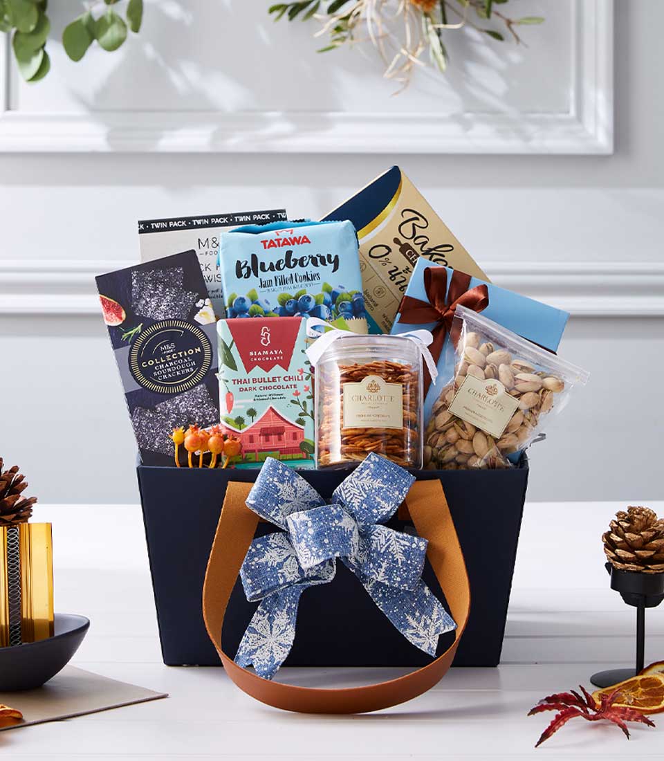 A luxurious Basketeer gift basket overflowing with gourmet indulgences, including decadent chocolates, delightful blueberry snacks, a jar of premium nuts, and a pouch of exquisite pistachios. The elegant basket is adorned with a lavish blue and white bow accented by the signature Basketeer ribbon, while charming pinecones enhance its festive allure. Set against a backdrop of sophisticated white paneling, this scene exudes refined elegance and inviting warmth.