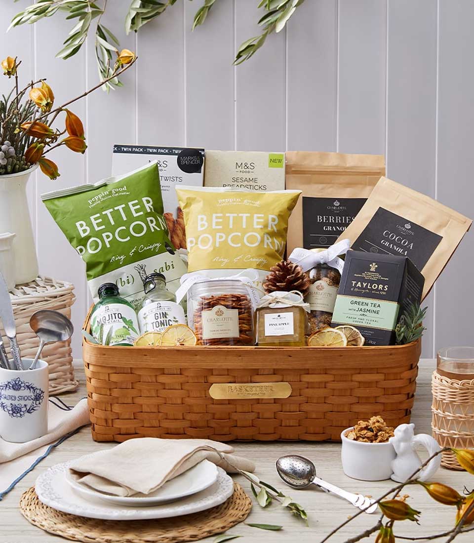 A luxurious wicker basket from Basketeer, adorned with a silky ribbon featuring the unmistakable Basketeer logo, elegantly graces a beautifully set dining table. The basket overflows with gourmet delights: artisanal popcorn, handcrafted breadsticks, premium crackers, exquisite chocolates, and a selection of fine teas. The table is invitingly arranged with an elegant cup and saucer beside refined plates and polished cutlery. Nearby sits a bowl of sumptuous cereal. Completing the scene is an eye-catching vase filled with fresh flowers, adding an extra touch of sophistication to this decadent display.