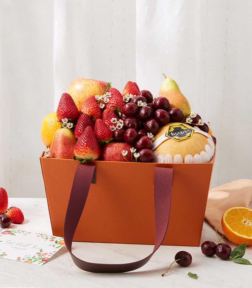 A luxurious orange basket, elegantly branded as the Basketeer Premium Fruit Temptations Gift, overflows with an exquisite selection of the finest fresh fruits. Succulent strawberries, luscious grapes, perfectly ripe pears and apples, and a sweet orange melon are nestled together in a mouthwatering display. Delicate white flowers thoughtfully adorn this tempting assortment. The basket itself features sophisticated brown handles, adding to its charm and opulence. Set against a pristine white surface, the scene is accentuated by a stylish card placed beside it—a perfect touch for any occasion deserving of elegance and indulgence.