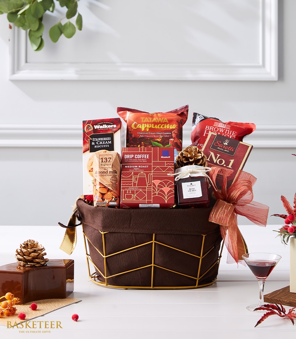 A luxurious festive gift basket from Basketeer, brimming with an exquisite assortment of gourmet delights including premium coffee, artisanal cookies, decadent brownie mix, and a jar of delectable jam. This opulent basket is adorned with elegant pine cones, vibrant red flowers, and a beautifully tied ribbon featuring the Basketeer logo. Beside the lavish arrangement is a crystal martini glass filled with a tantalizing cranberry cocktail, creating an inviting scene of holiday indulgence.