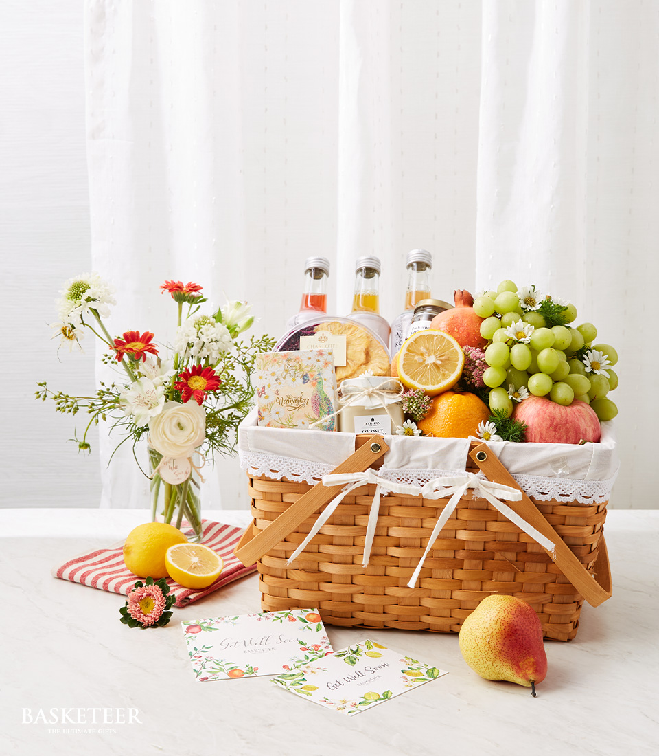 A Brown Rattan Gift Basket Filled with many types of fresh fruits such as red apples, green grapes, oranges, etc. and refreshing fruit juices. With fresh flowers in a bright, beautiful vase.
