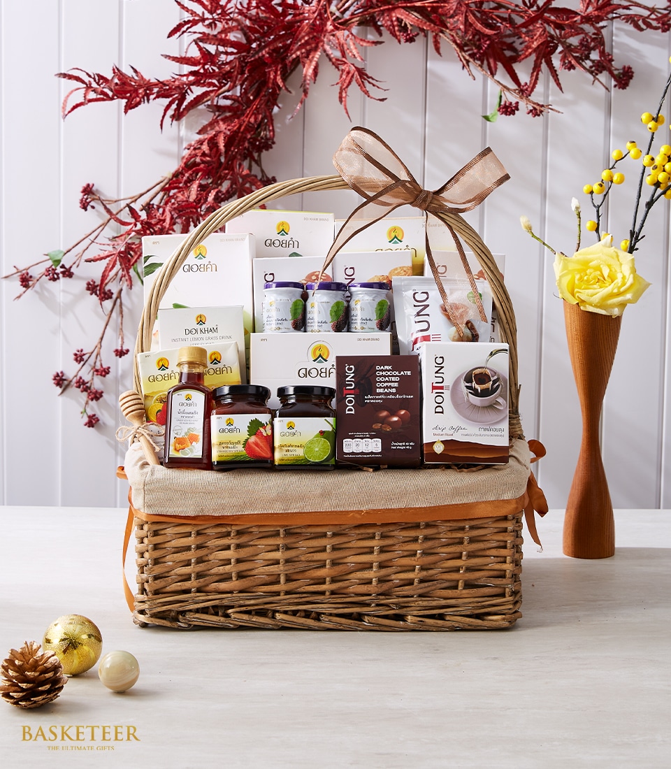 A luxurious wicker gift basket, elegantly tied with a rich brown ribbon featuring the Basketeer logo, overflows with an assortment of premium products encased in gorgeous jars, exquisite boxes, and sophisticated bottles. This stunning ensemble is complemented by a backdrop of vibrant autumnal red leaves and a delicate yellow rose decoration. In front, a charming pinecone and an opulent golden ornament add to the lavish ambiance.