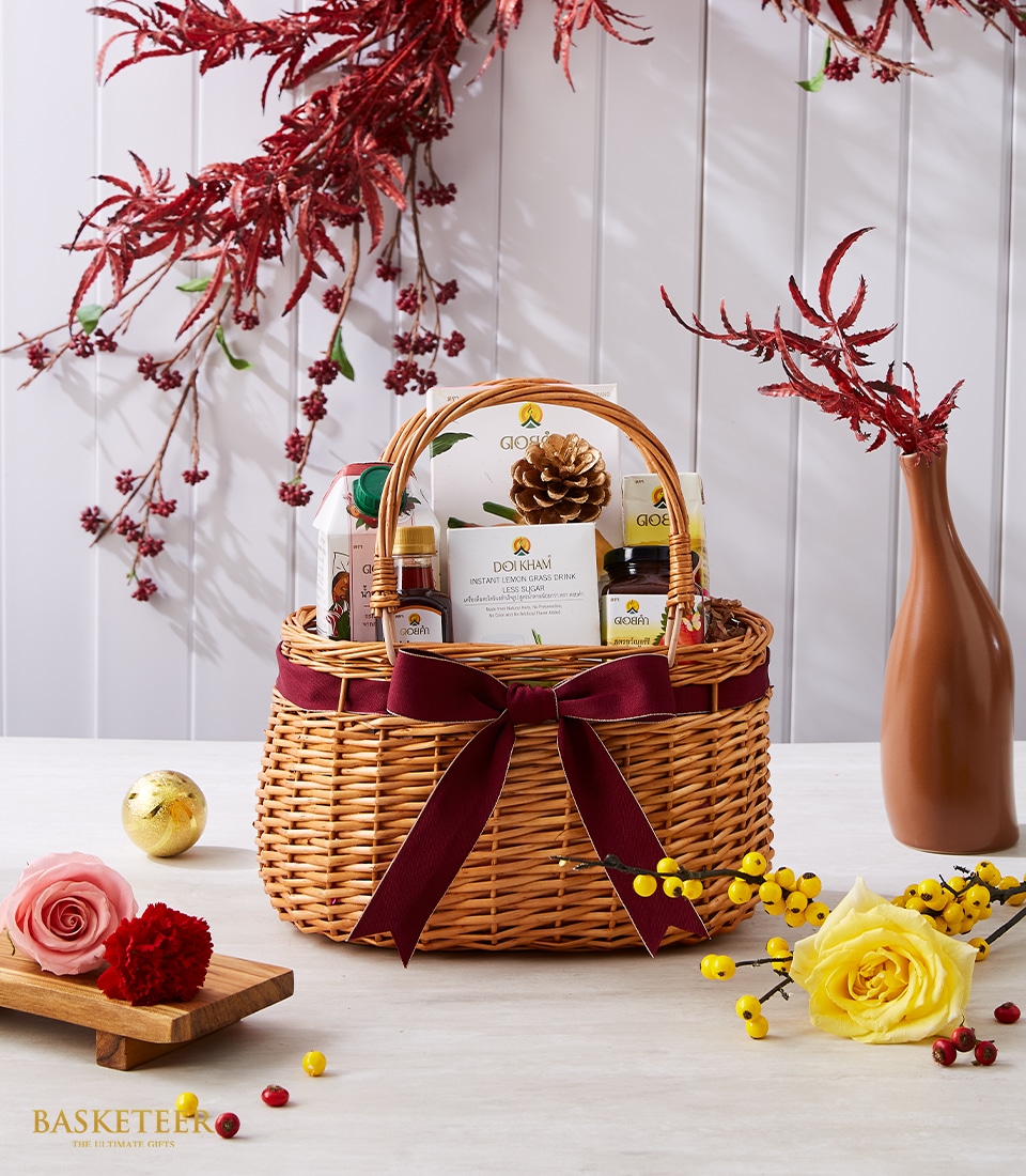 A luxurious wicker gift basket, elegantly adorned with a deep red bow featuring the Basketeer logo, brims with an exquisite selection of lotions and gourmet snacks. It graces a pristine white table, surrounded by decorative flowers and festive ornaments that enhance its opulent presentation. In the background, rich red foliage drapes gracefully, imbuing the scene with a warm holiday ambiance.