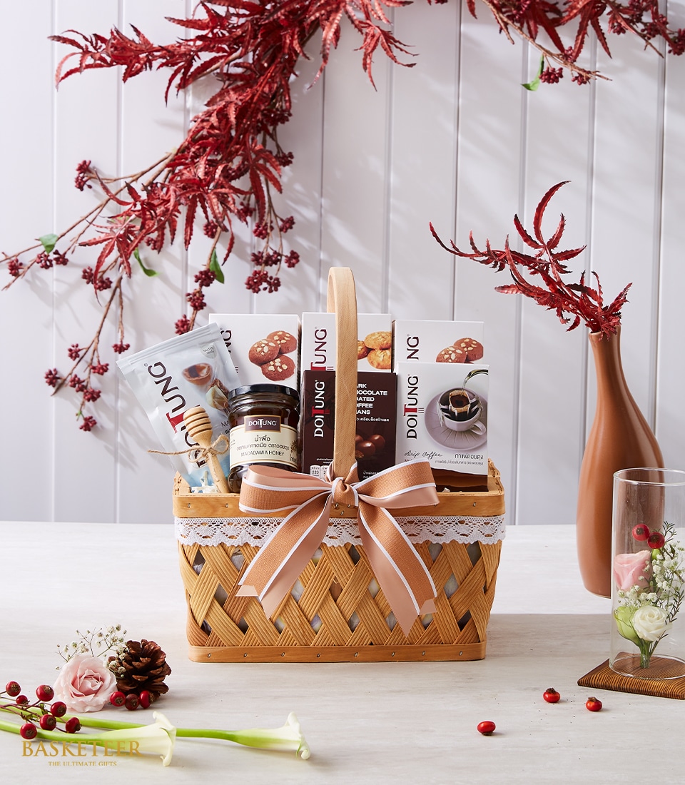 A luxurious wicker gift basket adorned with an elegant pink bow, proudly showcasing the Basketeer logo, is brimming with an exquisite selection of chocolate-covered nuts and gourmet spreads. This opulent arrangement rests on a pristine white table graced with vibrant red and pink flowers. In the backdrop, lush red foliage enhances a sophisticated white wooden panel, adding an enchanting ambiance to this sumptuous offering from Basketeer.