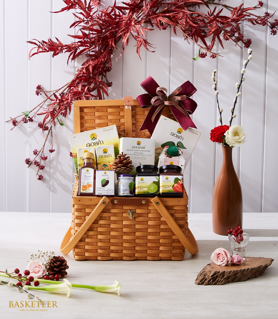 A luxurious wicker gift basket, curated by Basketeer, overflowing with gourmet snacks, freshly pressed juices, and artisanal spreads. The basket is elegantly adorned with a rich burgundy ribbon and bow featuring the Basketeer logo. It is beautifully set on a table surrounded by delicate floral decorations in a vase, against an inviting white and pink-themed background.