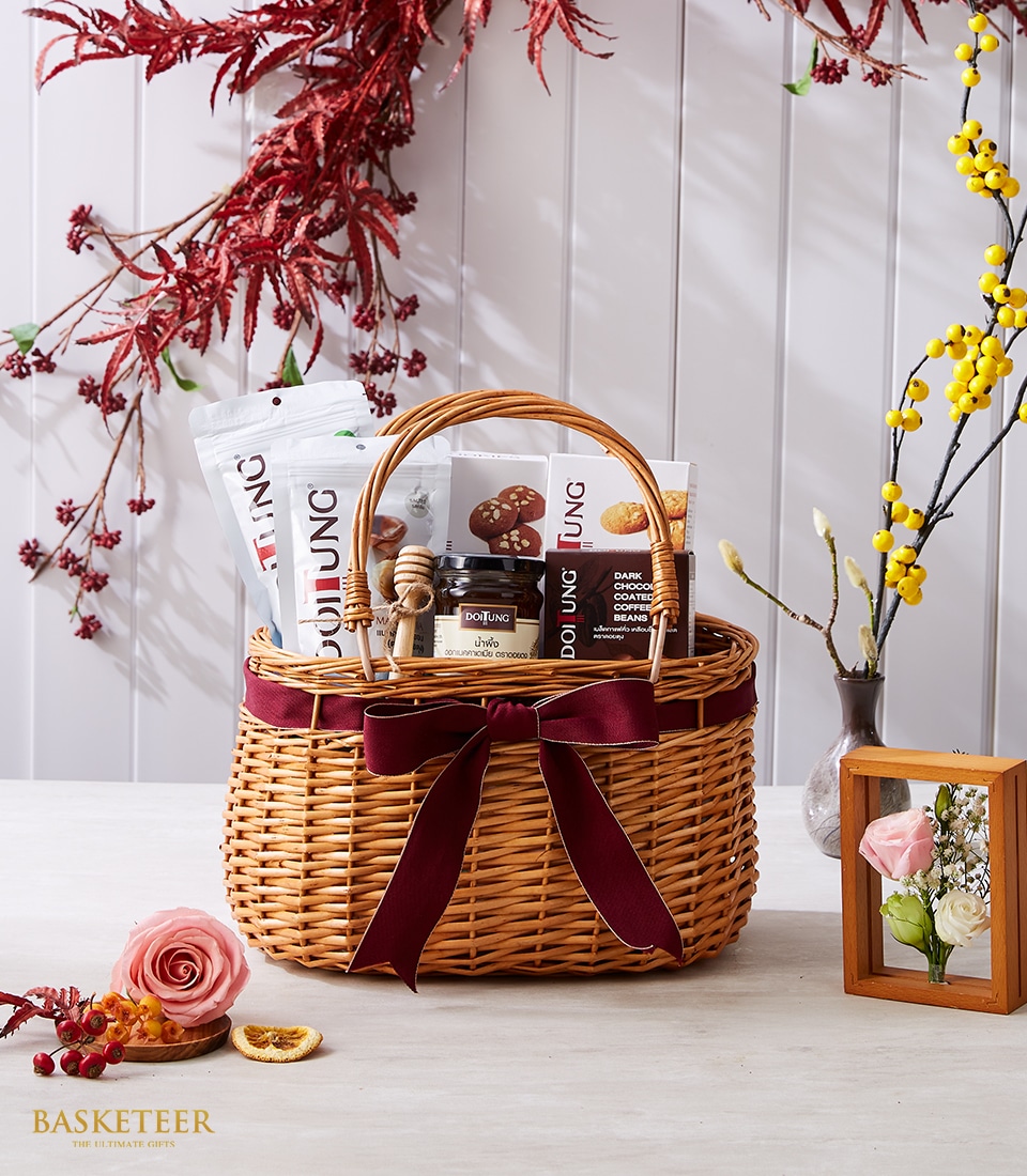 A lavish wicker basket graced with a sumptuous maroon ribbon and the Basketeer logo cradles an exquisite assortment of gourmet delights, such as artisanal honey dipper sticks, premium snacks, and curated dried goods. Elegantly positioned on an opulent table, this scene is accentuated by a delicate rosebud, a vintage vignette frame, and a sophisticated vase adorned with branches bearing berries and vibrant yellow blossoms—creating an inviting atmosphere of refined luxury.