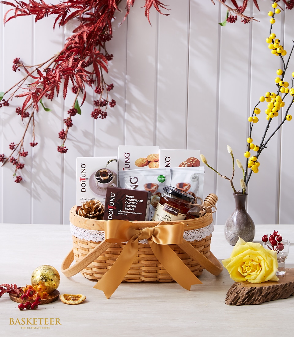 A luxurious wicker gift basket adorned with a shimmering gold ribbon proudly showcases the Basketeer logo. Inside, you'll discover indulgent Douling chocolates, an elegantly crafted jar, and decorative pinecones – all offering a touch of opulence. The backdrop is enriched with vibrant red foliage on the left and delicate yellow blossoms gracefully displayed in a chic vase on the right. In the foreground, a beautifully placed yellow rose and charming red berries enhance the scene's inviting allure.