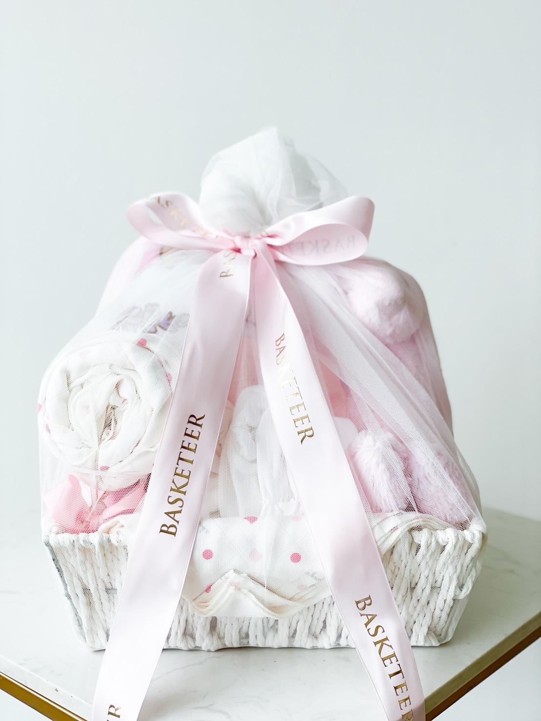A white basket wrapped in sheer white fabric, tied with a pink ribbon that reads 