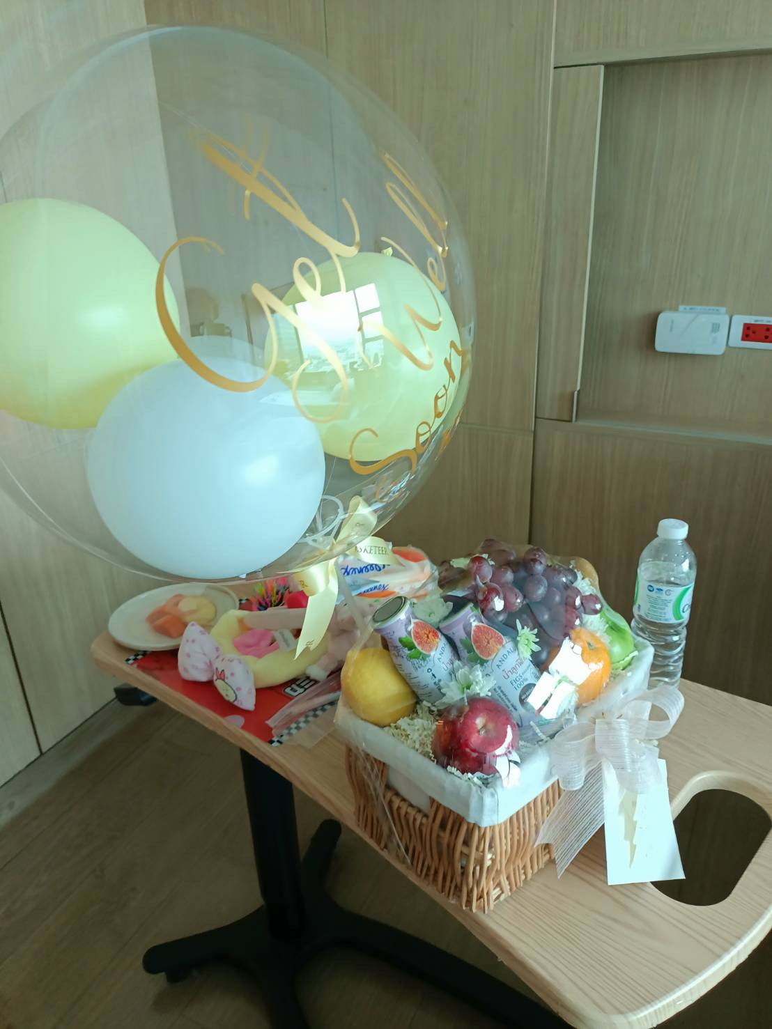 A cheerful gift arrangement featuring a wicker basket filled with fresh fruits like apples, grapes, and oranges, paired with a clear bubble balloon decorated with yellow and white accents, displayed on a table in a cozy indoor setting.