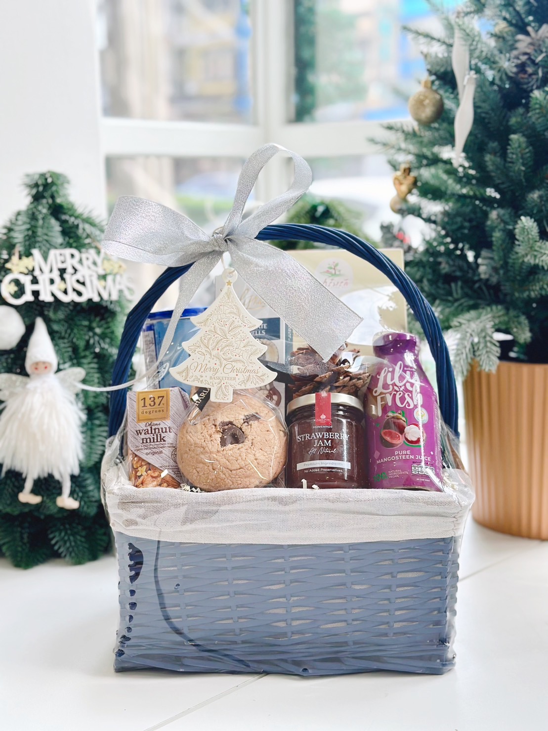 A beautifully arranged Christmas gift basket with a blue handle, decorated with a silver bow and a Christmas tree ornament, filled with premium goodies including cookies, jam, walnut milk, and mangosteen juice, placed in a festive setting with a Christmas tree and decorations in the background.