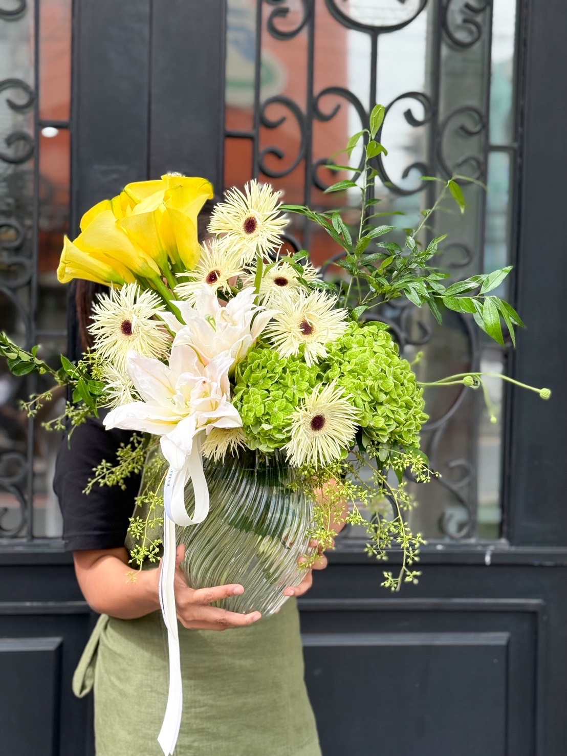 Elegant glass vase arrangement with yellow calla lilies, white lilies, green hydrangeas, and spider chrysanthemums, accented with lush greenery.