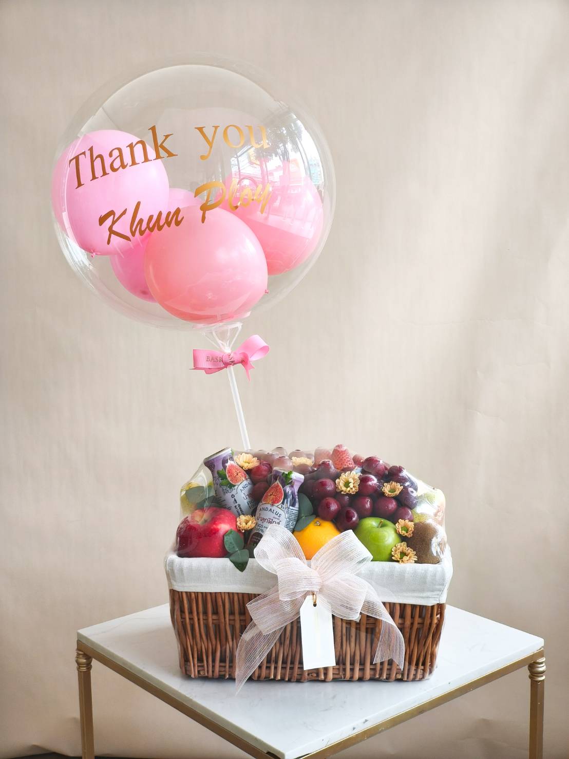 Luxury fruit basket with apples, grapes, oranges, pears, and strawberries, decorated with pink thank you balloon and satin ribbon.