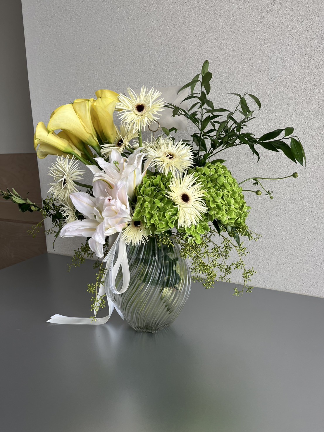 Elegant glass vase arrangement with yellow calla lilies, green hydrangeas, white lilies, cream gerberas, and lush foliage.