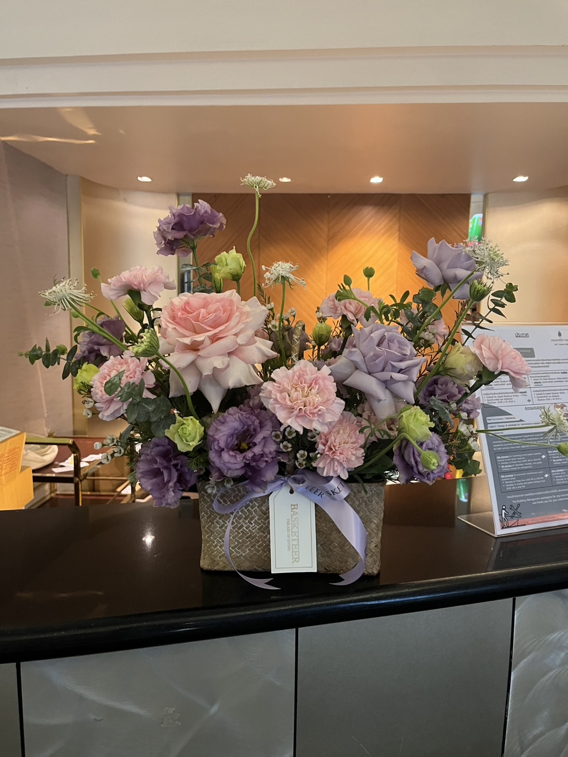 An Ocean Song Roses Basket filled with pink and purple blooms, including roses and carnations, sits on a counter. A lavender ribbon and gift tag add charm. The background reveals a warmly lit interior and a menu stand.