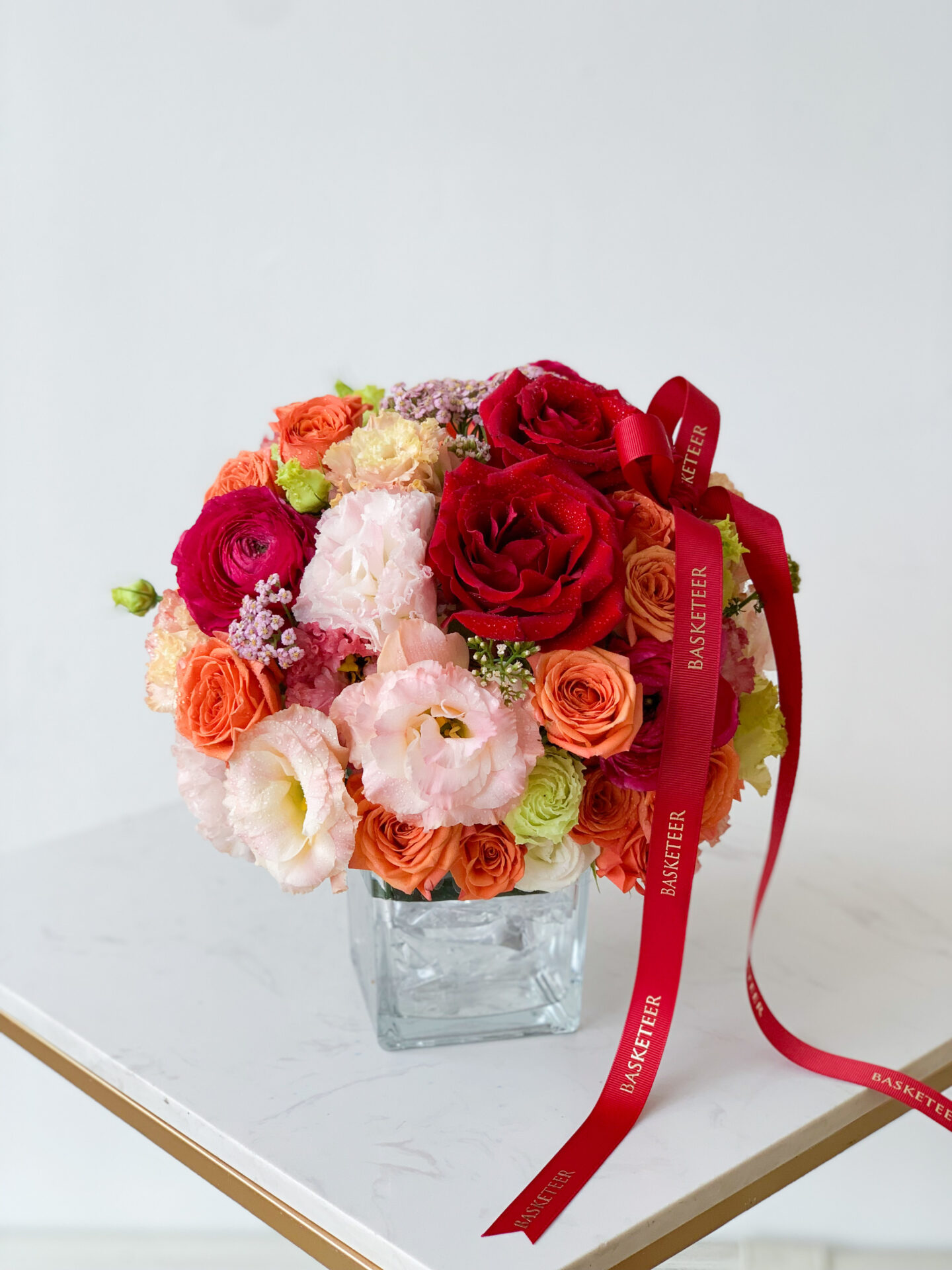 A glass vase is filled with a vibrant assortment of roses, peonies, and other flowers in shades of red, pink, peach, and green. A red ribbon with the word 