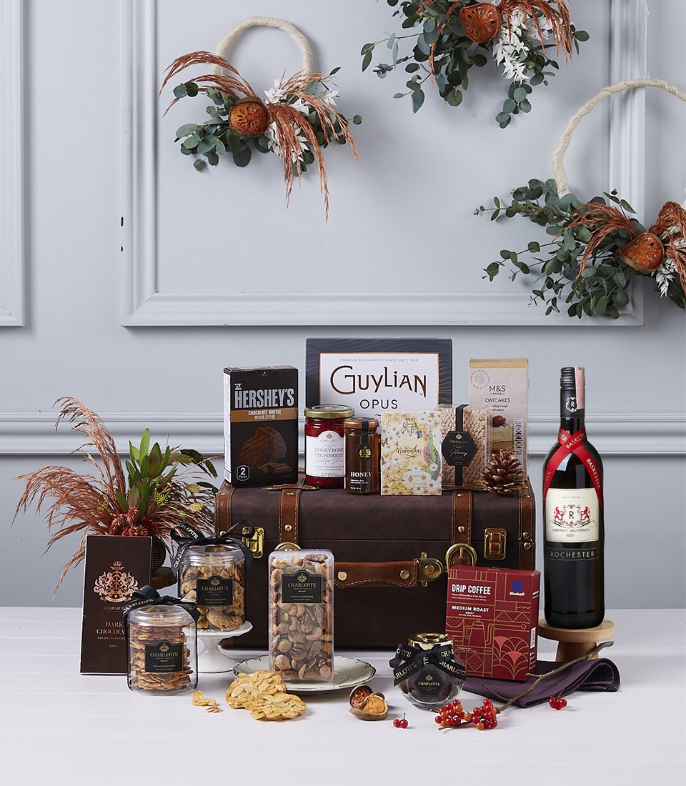 A luxurious gift hamper set on a white table features a variety of gourmet treats, including a bottle of red wine, chocolates, nuts, jam, crackers, and cookies. The backdrop includes decorative wreaths made of dried oranges, pine cones, and greenery.