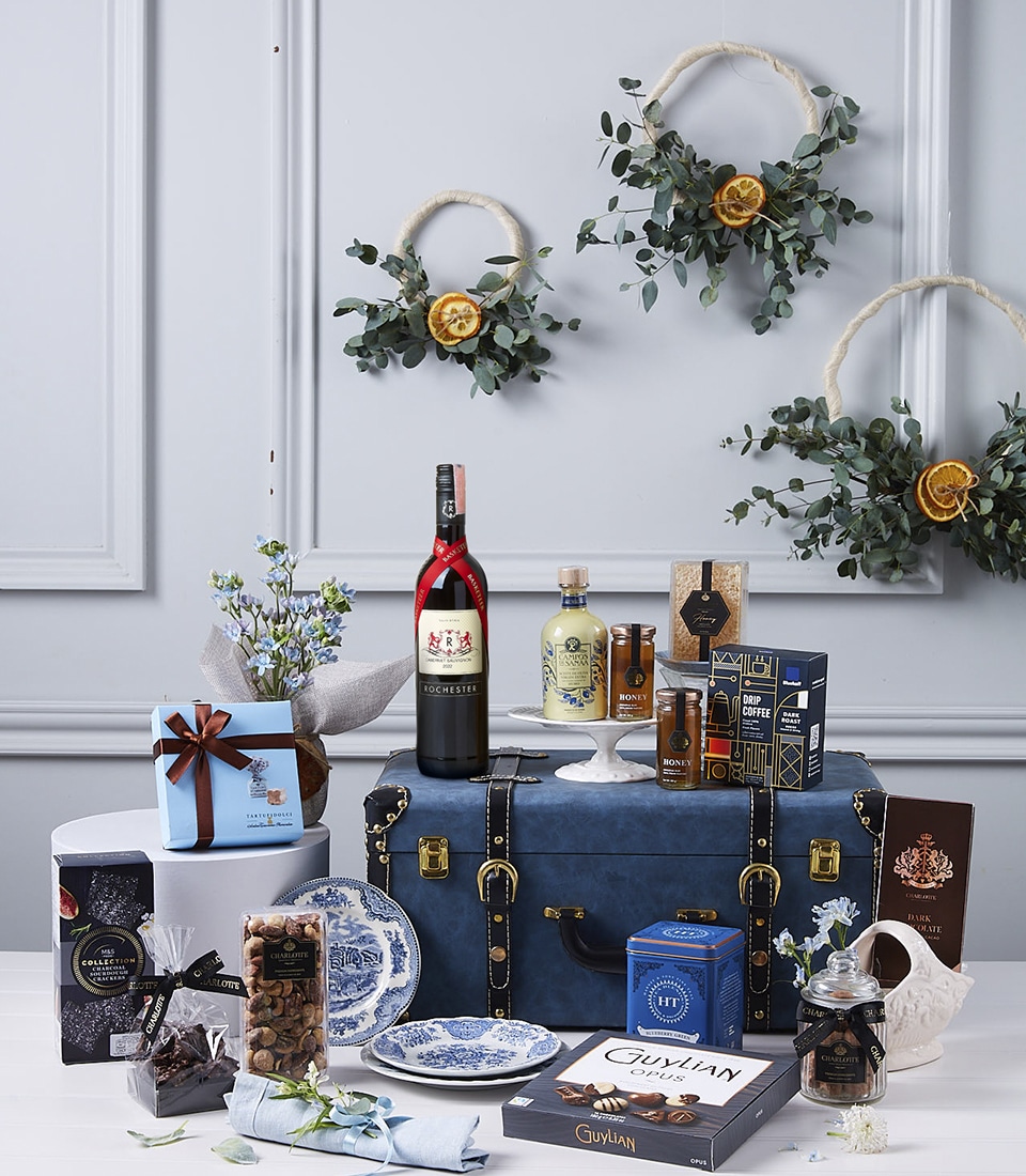A luxurious gift display featuring a bottle of red wine, chocolates, jams, and biscuits. These items are arranged around a dark blue trunk with gold accents. Decorative wreaths and dried citrus adorn the wall in the background, enhancing the elegant setup.