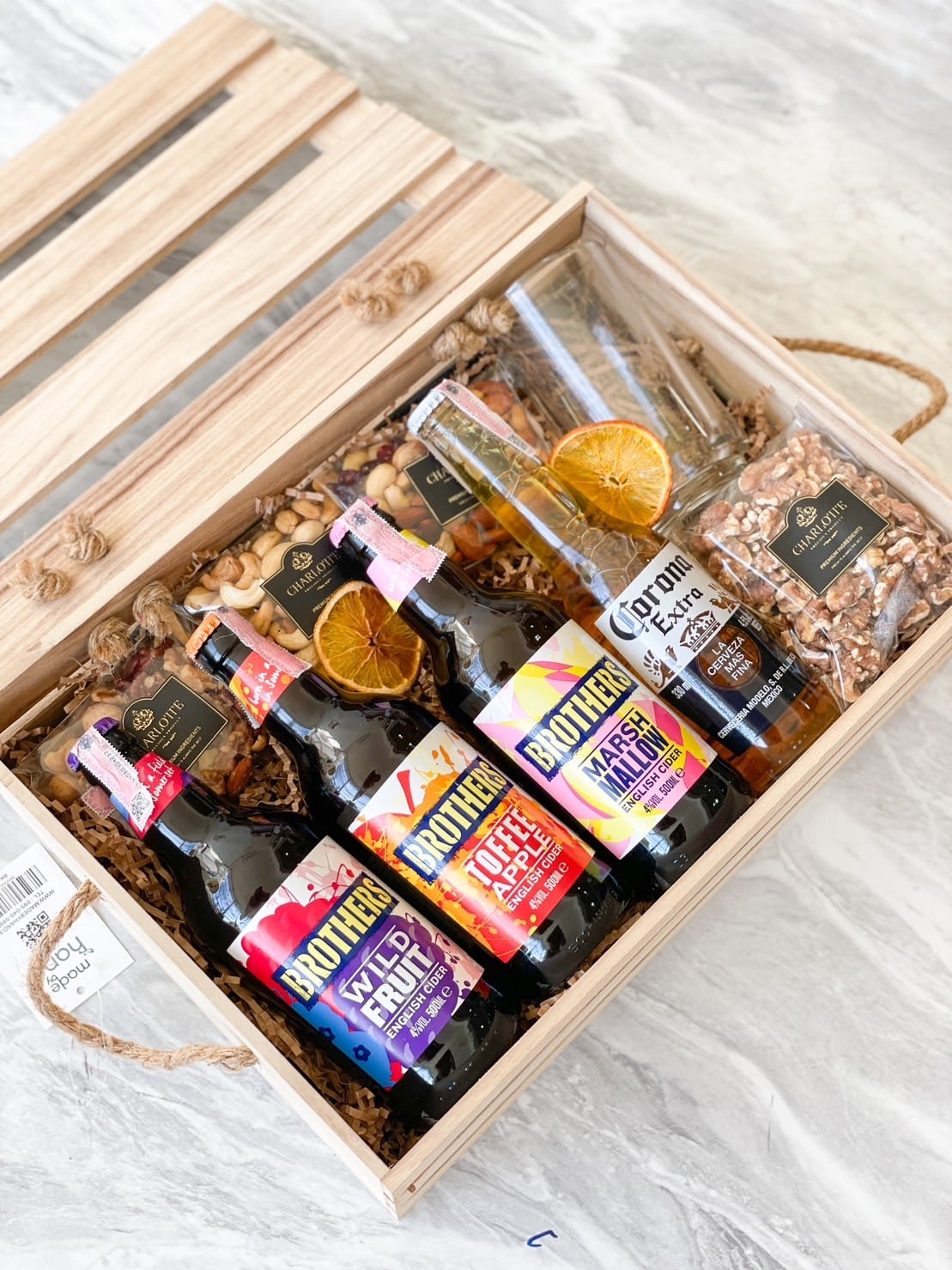 A wooden crate gift box contains a variety of items, including bottles of flavored beverages, a bottle of Corona Extra beer with a dried citrus garnish, and small bags of nuts. The crate features straw filling and has two handles made of braided rope.