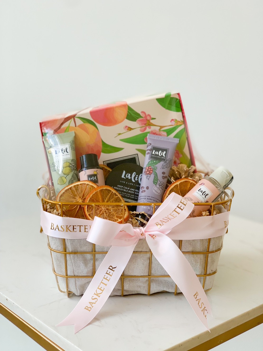 A Spa Relaxation Basket containing various personal care products including lotions, creams, and hand sanitizer, decorated with dried orange slices. The basket has a pink ribbon with