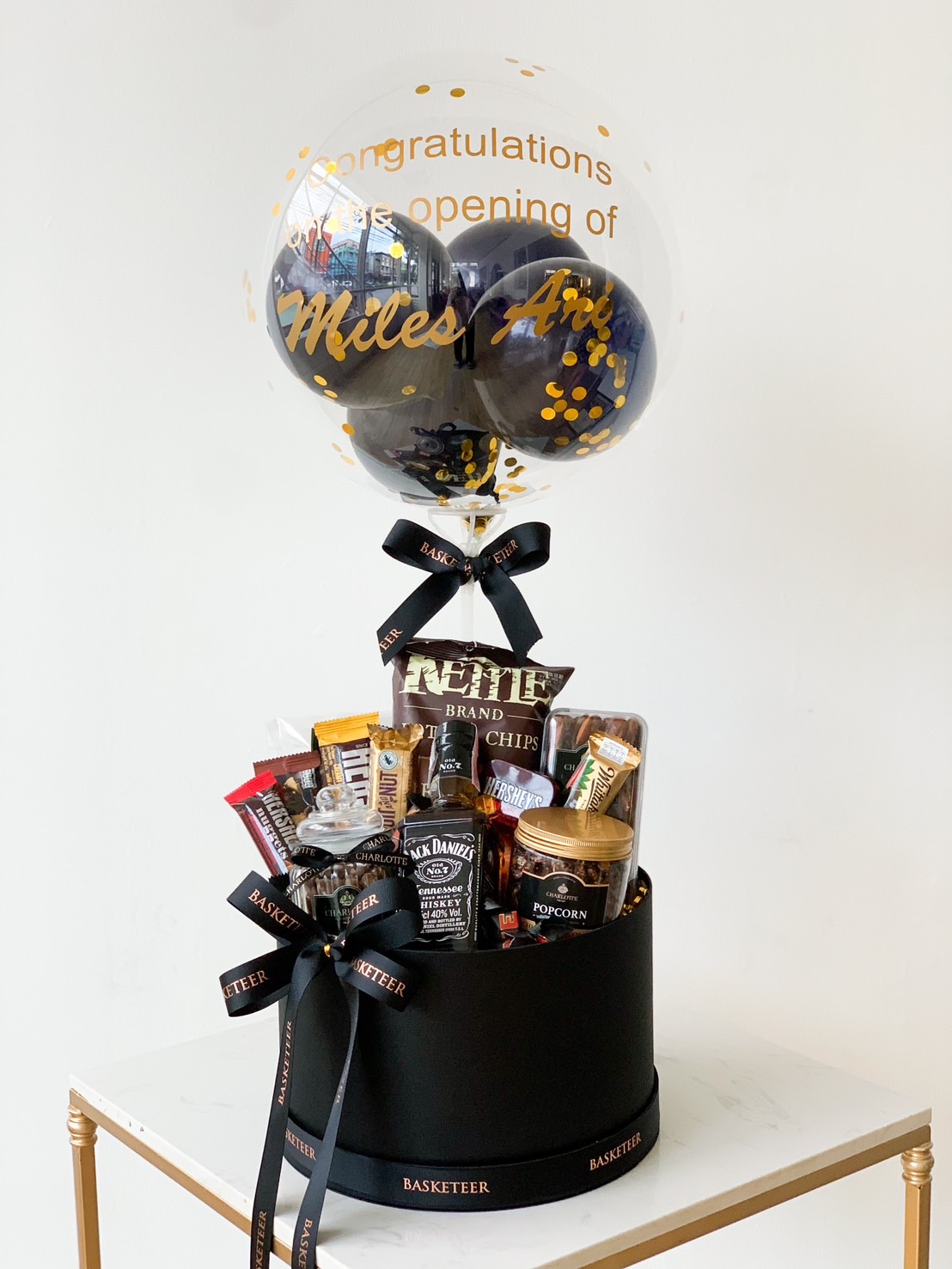 A black gift basket filled with snacks, including chips, popcorn, and Jack Daniel's products, topped with a clear balloon containing black and gold confetti. The balloon has a congratulatory message for the opening of "Miles Inc." written in gold.