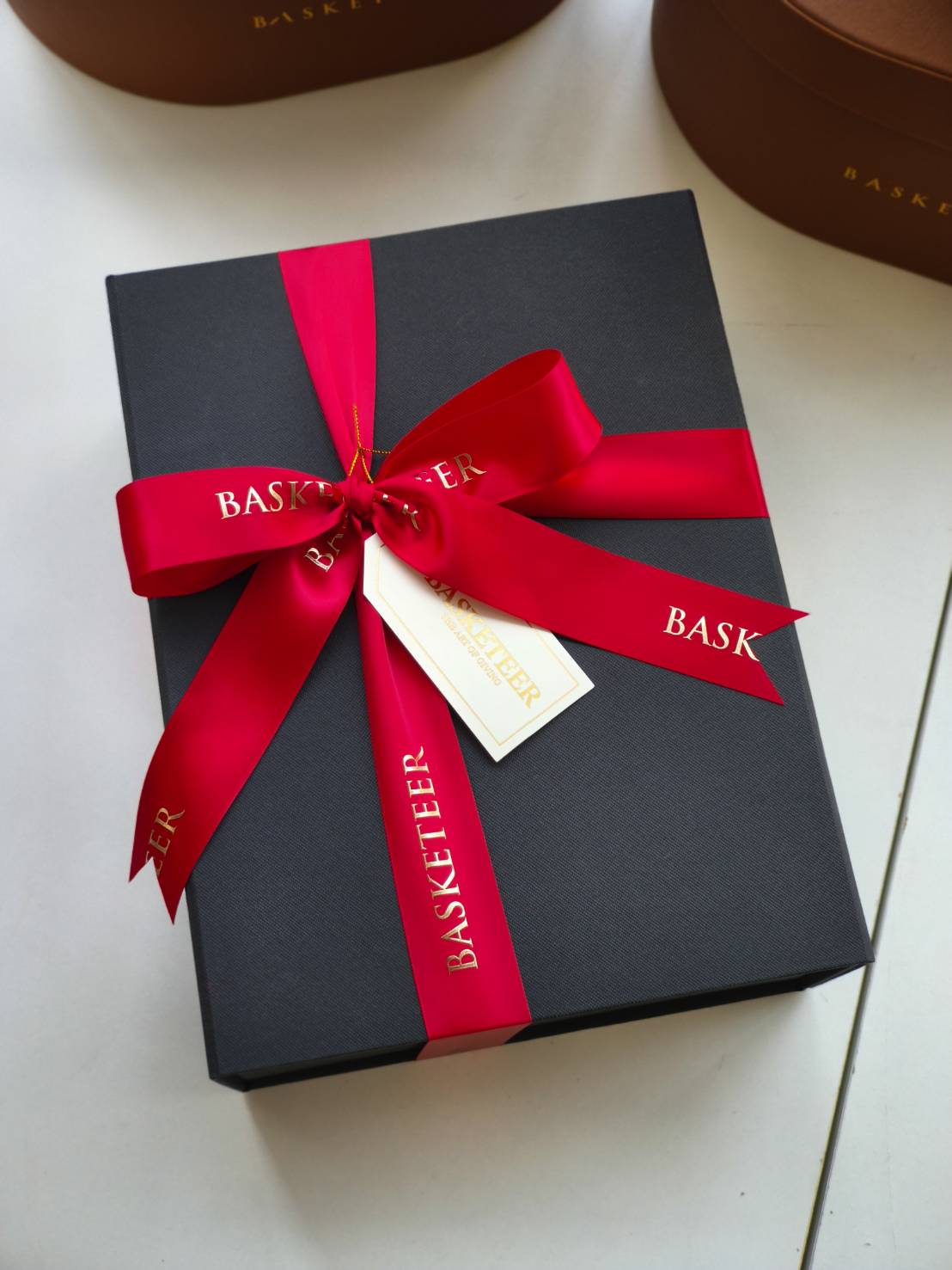 Elegant black gift box tied with a wide red satin ribbon printed with “Basketeer” in gold lettering.