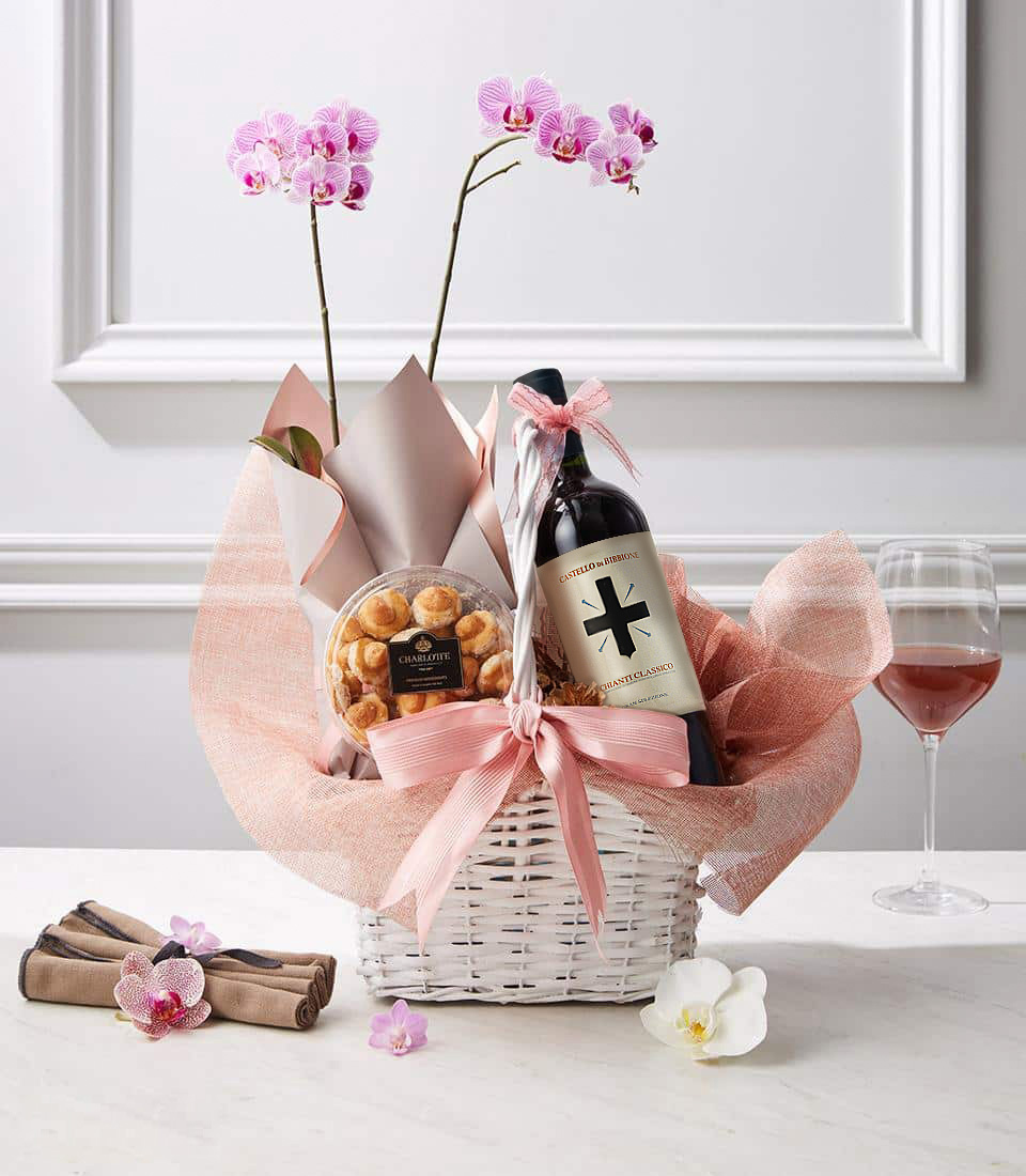 A sophisticated gift basket featuring a bottle of red wine, a delicate orchid plant, and gourmet pastries, elegantly wrapped in pink fabric with a satin bow.