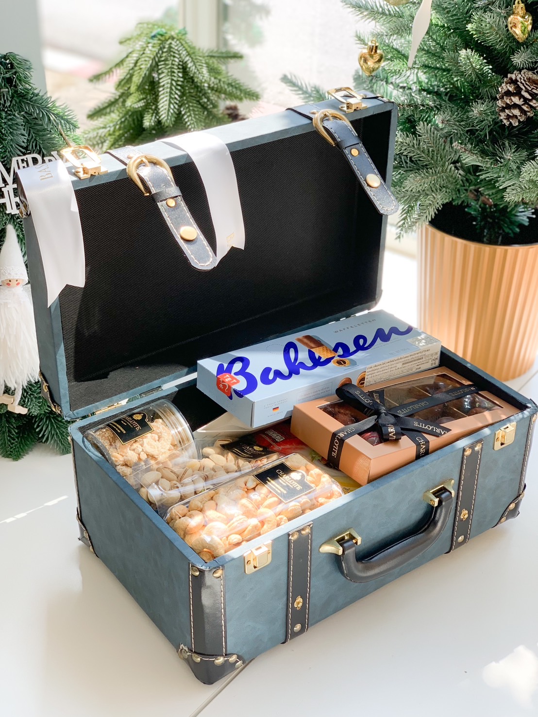 Open vintage blue leather trunk filled with gourmet treats, including chocolates, nuts, and pastries, against a festive Christmas background.