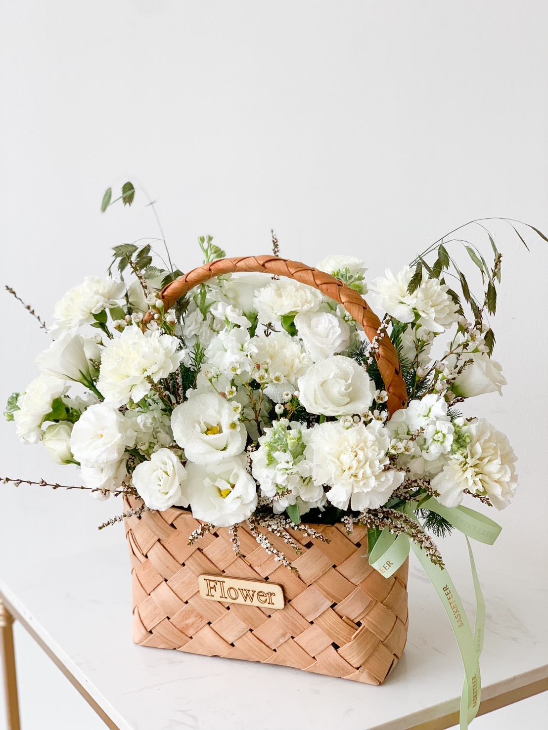 A wicker basket filled with an arrangement of white flowers, including roses and carnations, accented with green foliage. A green ribbon is tied around the handle of the basket, which has a small wooden plaque reading 
