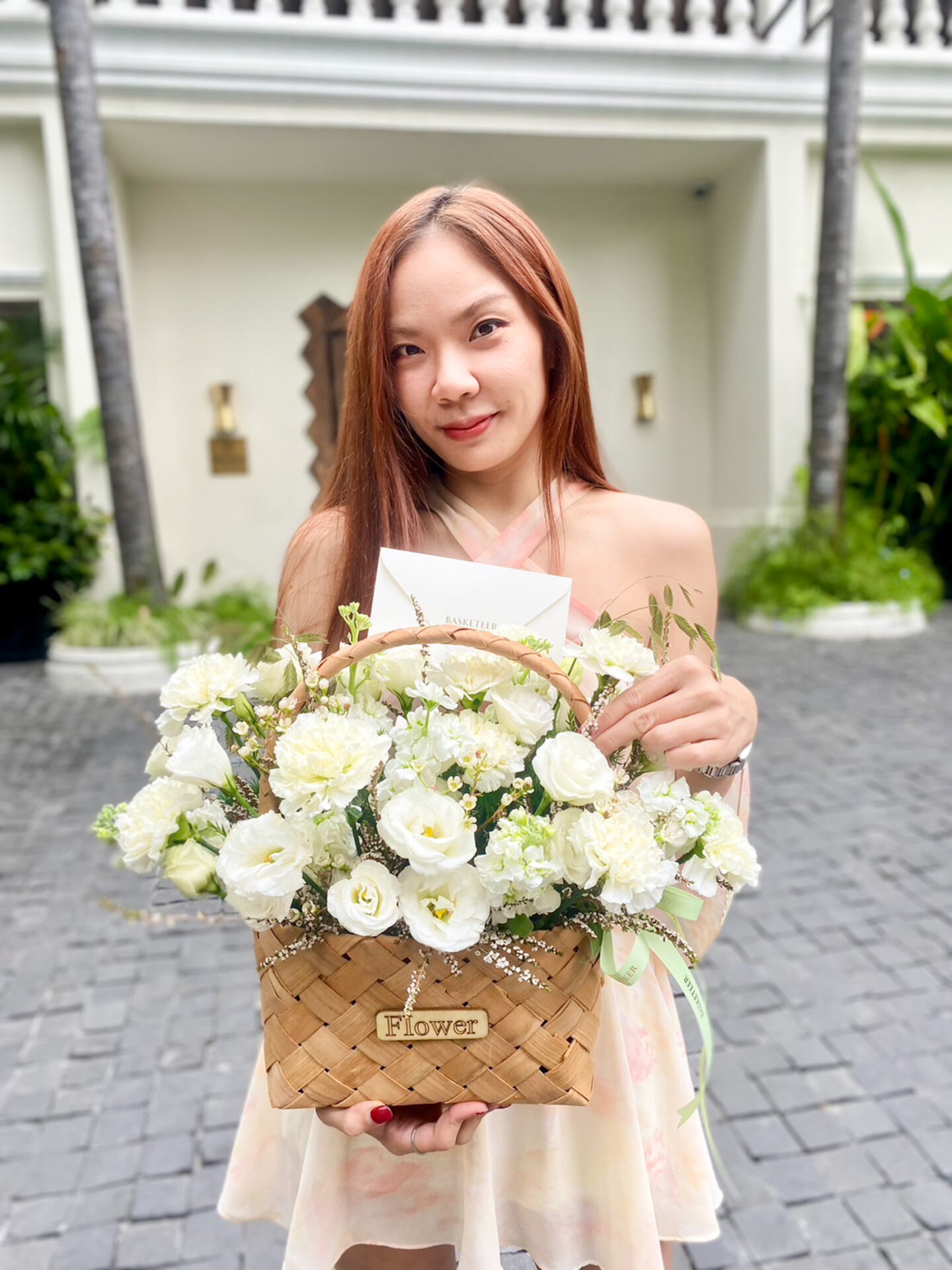 A woman with long hair stands outdoors in front of a white building surrounded by greenery. She holds a wicker basket filled with white flowers and greenery, with a card tucked among the blooms. She is wearing a light-colored dress and smiling at the camera.
