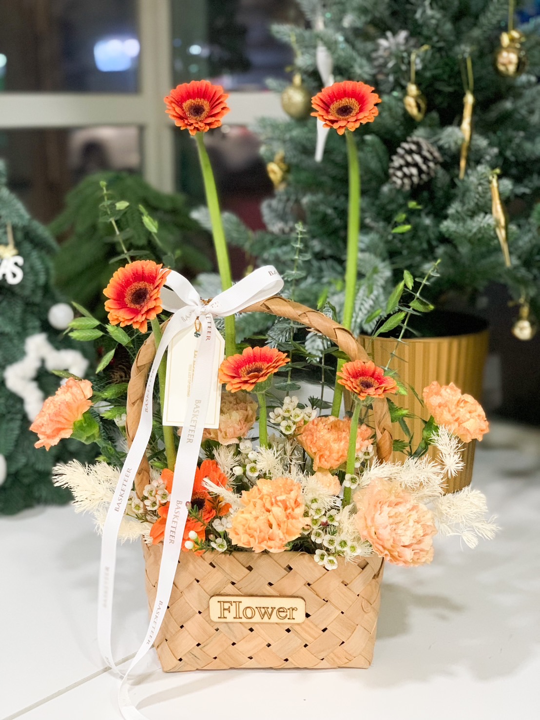 Basket of vibrant orange gerberas, peach carnations, and delicate white blooms, accented with greenery and a 