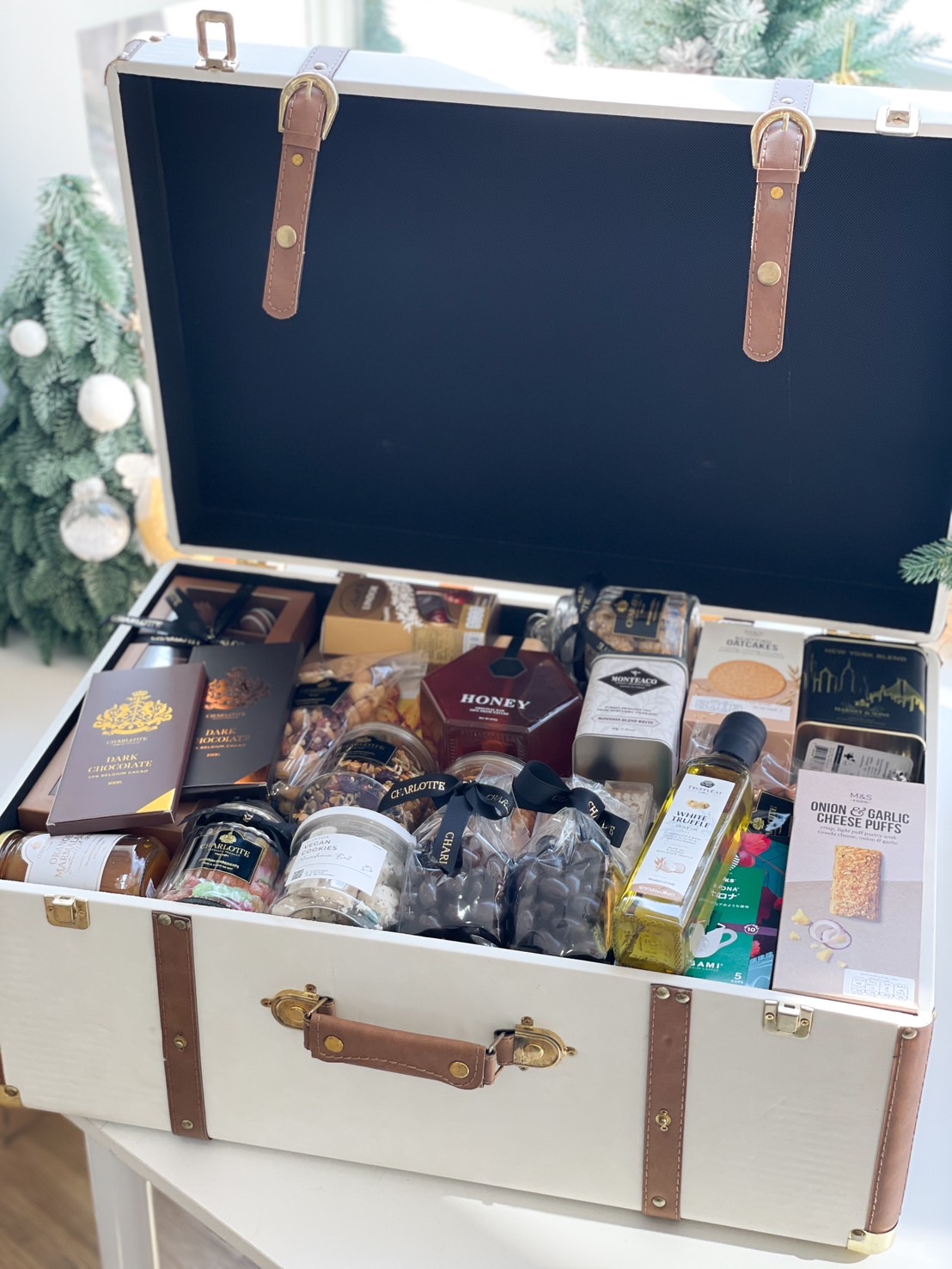 A white luxury trunk with leather accents, filled with gourmet treats including honey, dark chocolate, nuts, olive oil, and cheese puffs.