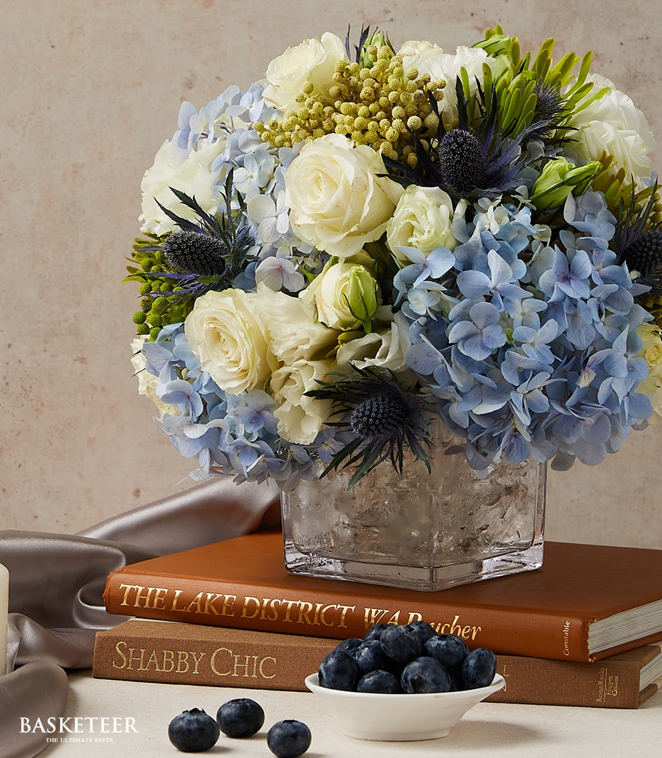 A lavish square glass vase cradles an exquisite arrangement of pristine white roses, delicate pale blue hydrangeas, and lush green foliage. This elegant display from Basketeer graces a refined surface, accompanied by two stylish books, 
