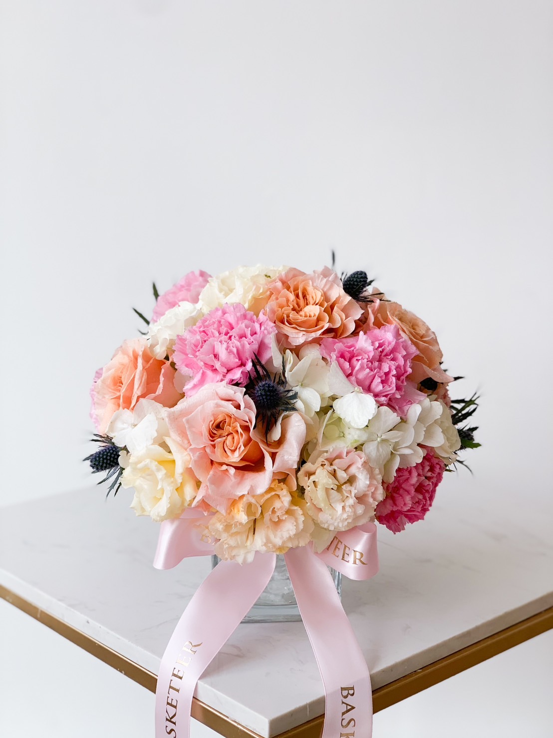 A round floral arrangement featuring pink, white, and peach roses with accents of dark thistle flowers, set in a clear vase on a white marble surface. The arrangement is adorned with a pink ribbon that has gold lettering.