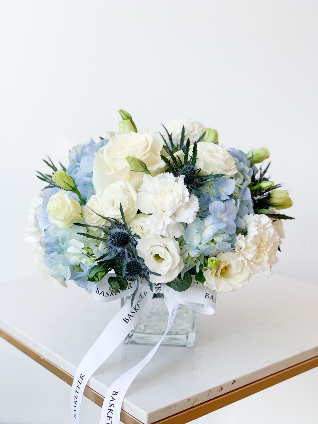 A floral arrangement featuring white roses, carnations, and lisianthus alongside light blue hydrangeas and thistles. The bouquet is in a clear glass vase on a white marble surface, adorned with white ribbon that has 