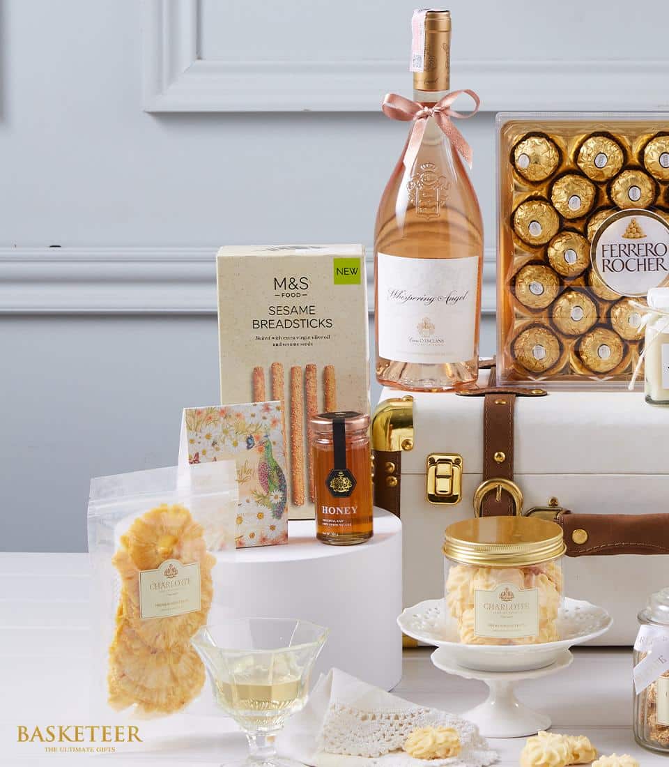 An exquisite gourmet gift set designed for indulgence, featuring a fine bottle of rosé wine, an elegant box of Ferrero Rocher chocolates, premium honey in a glass jar, crunchy sesame breadsticks, deliciously dried pineapples, and a charming jar of assorted sweets. These luxurious treats are meticulously arranged with care next to a pristine white suitcase and accompanied by a beautifully branded Basketeer card in the background – an invitation to savor life's finest moments.