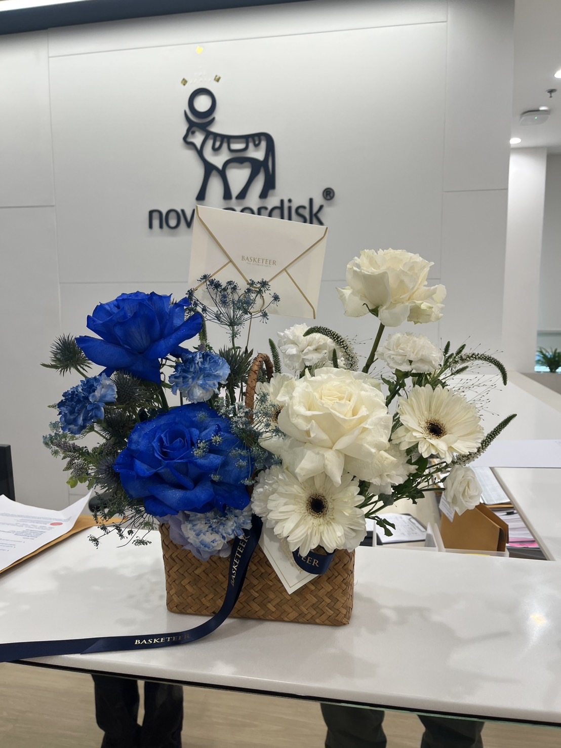 Elegant woven basket filled with blue roses, white roses, gerberas, hydrangeas, and thistles, tied with a navy ribbon.